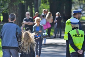 Policjanci ruchu drogowego nadzorujący przyszkolne przejście dla pieszych. W tle mężczyzna prowadzący dziewczynkę za rękę oraz inni przechodnie