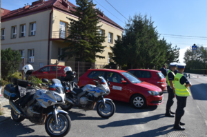 Budynek szkoły, obok policjanci w kamizelkach odblaskowych z napisem POLICJA. Policyjne motocykle oraz samochód z logo Małopolskiego Ośrodka Ruchu Drogowego w Tarnowie