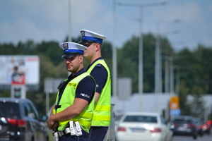 Dwóch poloicjantów ruchu drogowego stoi przy drodze nadzoruje ruch. W tle ruch uliczny słoneczna letnie pogoda