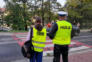 policjant ruchu drogowego w kamizelce odblaskowej stojący przy przejściu dla pieszych obok niego uczennica w kamizelce odblaskowej opisanej bezpieczny powiat krakowski