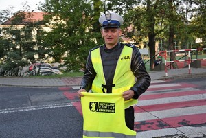 policjant trzymający w ręku kamizelkę odblaskową z napisem bezpieczny powiat krakowski