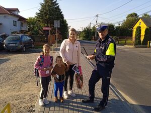 Policjant Ruchu Drogowego stoi na chodniku z natka z dwójką dzieci chłopczykiem i dziewczynką wręcza im elementy odblaskowe w tle parking samochodowy, zabudowania i ulica słoneczna pogoda