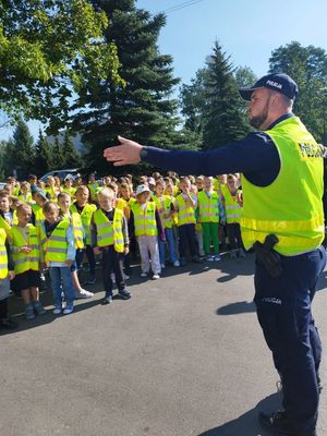 Policjant w mundurze w kamizelce odblaskowej stoi twarzą do grupy uczniów, krórzy rownież mają na sobie kamizelki odblaskowe. W tle widać drzewa i słoneczą pogodę.