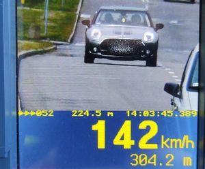 Zdjęcie ręcznego miernika prędkości, na którego wyświetlaczu jest srebrny samochód Mini, który porusza się w dzień po asfaltowej drodze. Poniżej wyświetlona jest zmierzona prędkość 142 kmh oraz odległości