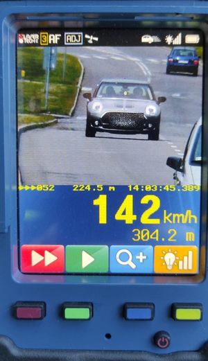 Zdjęcie ręcznego miernika prędkości, na którego wyświetlaczu jest srebrny samochód Mini, który porusza się w dzień po asfaltowej drodze. Poniżej wyświetlona jest zmierzona prędkość 142 kmh oraz odległości