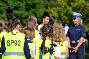 Dzieci w kamizelkach odblaskowych z napisem SP92 słuchają prelekcji w terenie prowadzonej przez policjantkę ruchu drogowego