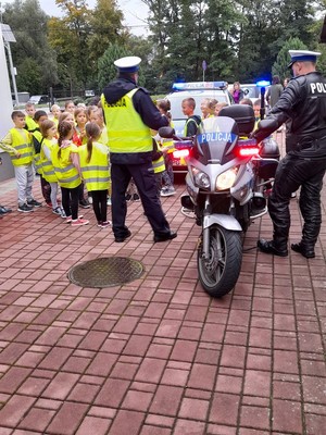 Policjanci oraz dzieci w kamizelkach odblaskowych, stojące obok policyjnego radiowozu i motocykla w których uruchomione są sygnały świetlne.