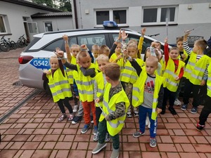 Dzieci w kamizelkach odblaskowych, podniosły do góry ręce na których mają założone elementy odblaskowe. Dzieci stoją obok policyjnego radiowozu.