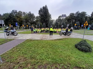 Dwa policyjne motocykle, umundurowani policjanci oraz dzieci w kamizelkach odblaskowych i nauczyciele.