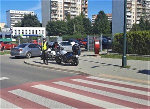 oznakowane przejście dla pieszych przy którym stoi umundurowny policjant  obok motocykl służbowy, w tle samochody i budynki