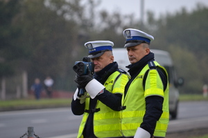 Dwóch policjantów ruchu drogowego (białe czapki, żółte kamizelki) zandzorują ruch na drodze. Jeden z nich posługuje się ręcznym miernikiem prędkości pojazdów.