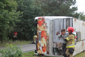 strażak prowadzący poszkodowanego w wypadku mężczyznę, w tle przewrócony na bok autobus