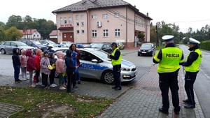 Policjanci i grupa dzieci stoją przy radiowozie. Policjant wyjaśnia zasady poruszania się po drodze