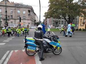 Grupa dzieci w kamizelkach odblaskowych idzie ulicą w towarzystwie opiekunów i maskotek różnych instytucji. Na pierwszym planie policjant na służbowym motocyklu