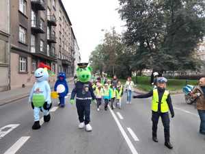 Policjantka, maskotki instytucji oraz dzieci w kamizelkach odblaskowych maszerują ulicą