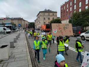 Uczniowie w kamizelkach odblaskowych niosą transparent i przechodzą jedną z krakowskich ulic