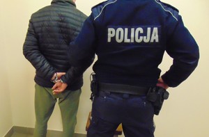 Zatrzymany w kajdankach stoi przy ścianie za nim umundurowany policjant