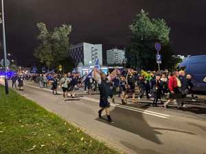 Liczna grupa kibiców z flagami, ubrana w szkockie stroje zierza w kierunku stadionu