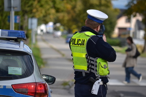 Policjant ruchu drogowego kontroluje ręcznym miernikiem prędkość pojazdów. W tle przejście dla pieszych i przechodząca przez nie kobieta