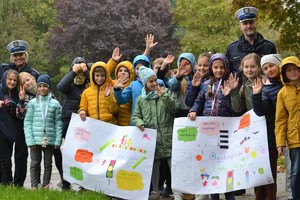 Grupa uczniów szkoły podstawowej stojąca na zewnątrz w towarzystwie policjantów ruchu drogowego prezentująca transparenty z hasłami promującymi noszenie odblasków