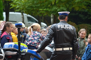 Policjant ruchu drogowego ubrany w skórzany mundur motocyklowy stoi przy mototrze otoczonym dziećmi. Jedno z dizeci siedzi na motorze pod okiem policjanta