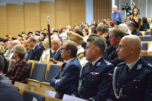 Komendant Miejski policji w Krakowie w auli uczelni słucha przemówienia Rektora uczelni, wokół inne osoby siedzące na krzesłach