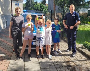dzielnicowy Jakub Ciągło  przy radiowozie obok nauczycielka i uczniowie z odblaskami w rękach