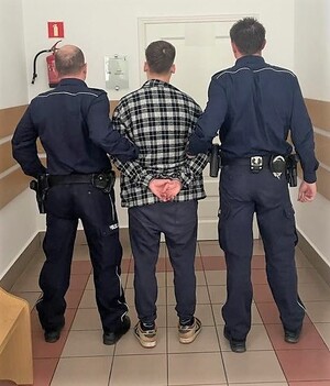 Zatrzymany mężczyzna, stojący tyłem. Ubrany w ciemne spodnie i koszulę w kratkę. Na rękach trzymanych z tyłu ma kajdanki. Po prawej i po lewej stronie stoją umundurowani policjanci, tyłem, którzy go trzymają.