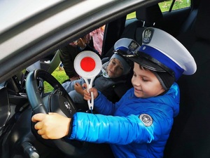 dziecko siedzi za kierownicą samochodu służbowego na głowie ma czapkę ruchu drogowego a w ręku tarczę do zatrzymywania pojazdów, w tle widać inne dziecko