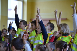 Grupa uczniów ubranych w kamizelki odblaskowe aktywnie bierze udział w prelekcji - widać mnogość podniesionych rąk