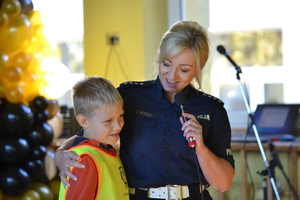 Policjantka ruchu drogowego trzyma w ręku mikrofon, drugą ręką obejmuje ucznia ubranego w kamizelkę, prowadzi z nim rozmowę