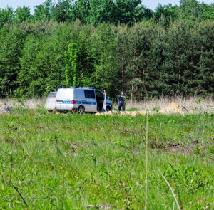 policjant stojący obrócony tyłem obok radiowozu w pobliżu kompleksu leśnego