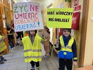 dwójka dzieci w kamizelkach odblaskowych na szkolnym korytarzu pokazuje transparenty promujące noszenie odblasków