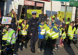 dzieci ubrane w kamizelki odblaskowe stoją przed budynkiem w towarzystwie policjantki
