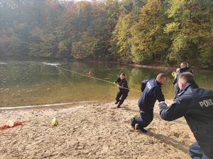 Trzech umundurowanych policjantów przy pomocy liny wyciąga na brzeg osobę,  znajdująca się w wodzie. Postawa policjantów w ruchu, akcja dynamiczna.   Obok po prawej stronie stoją komendanci. W tle widać drzewa. Pogoda jesienna.