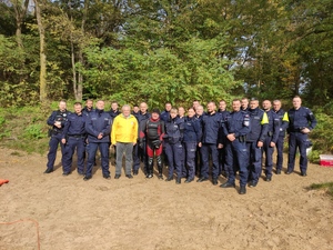Grupa policjantów oraz prezes i ratownik WOPR-u pozują do zdjęcia. W tle drzewa. Pogoda jesienna słoneczna.