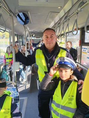 Umundurowany policjant ruchu drogowego w autobusie komunikacji miejskiej wraz z uczniem. Obaj maja kamizelki odblaskowe
