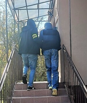 Zatrzymany mężczyzna. Ubrany w niebieskie dżinsy i kurtkę koloru czarnego, włosy krótkie ciemne. Idący po schodach. Obok niego policjant w stroju cywilnym. Na prawej ręce na opaskę z napisem Policja. Osoby są tyłem.