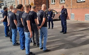 Parking przed budynkiem Komendy Miejskiej Policji w Krakowie. Rezerwiści w koszulkach z napisem Egida-22 stoją w dwuszeregi, przed nimi stoją dwaj policjanci