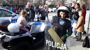 Młody chłopiec stoi ubrany w policyjny kask, a w rękach trzyma policyjną tarczę. Za nim na quadzie siedzi mała dziewczynka. Obok inne osoby oglądają policyjny sprzęt