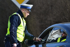 Policjant ruchu drogowego stoi przy samochodzie osobowym i bada siedzącego w środku kierowcę urządzeniem elektronicznym na zawartość alkoholu w wydychanym powietrzu