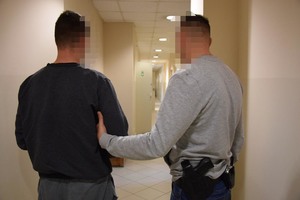 Policjant prowadzi po korytarzu zatrzymanego mężczyznę zakutego w kajdanki