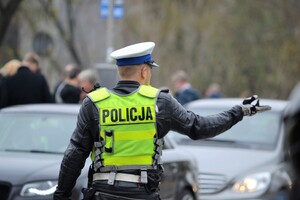Policjant ruchu drogowego ubrany w służbowy strój motocyklowy wśród zwmożonego ruchu wydaje gestami rąk polecenia w celu upłynnienia ruchu
