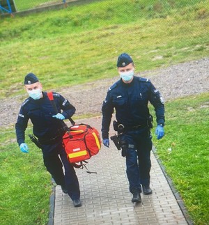 dwóch idących policjantów, jeden z nich trzyma na ramieniu czerwoną torbę ratowniczą