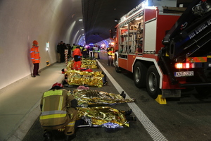 strażacy udzuelając pomocy rannym leżącym na ziemi owiniętym w koce termiczne. obok nic zaparkowany wóz strażacki