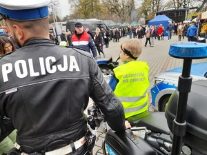 chłopiec w kamizelce odblaskowej siedzący na motocyklu policyjnym, obok policjant stojący tyłem, w tle grupa osób