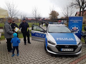 policjant stojący przy radiowozie wręczający kamizelkę odblaskową mężczyźnie z dziećmi, w tle uczestnicy pikniku oglądający mural