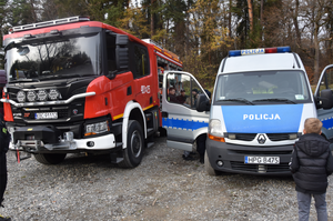 Osoba biorąca udział w akcji ubrana w elementy polciyjnego umundurowania. W tle zaparkowane pojazdy i inni uczestnicy akcji