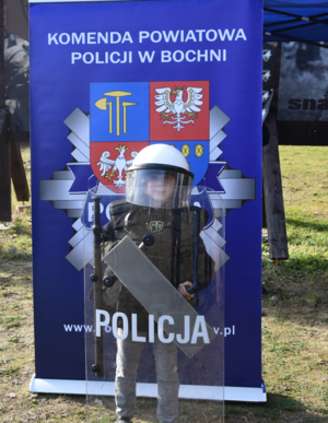 Uczestnik akcji ubrany w elementy policyjnego umundurowania, trzymający w rękach tarczę z napisem POLICJA, stojący na tle baneru z logo Komendy Powiatowej Policji w Bochni