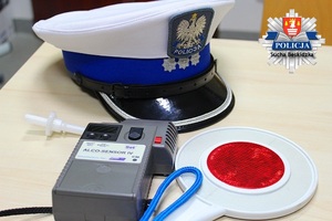 Położona na stoliku czapka funkcjonariusza ruchu drogowego z białym otokiem a obok tarcza do zatrzymywania samochodow oraz urządzenie do badania